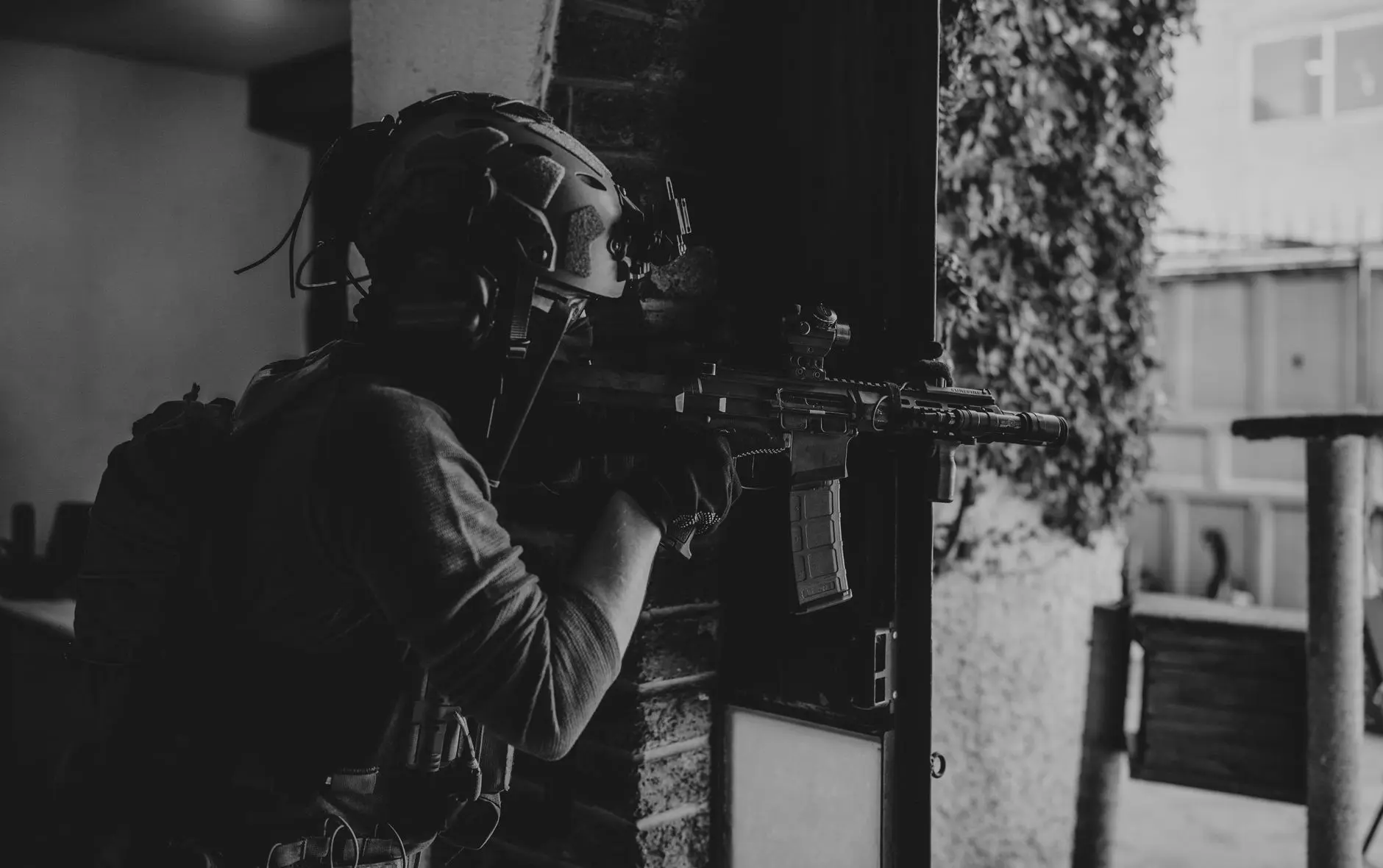 Black and white photo of a tactical operator in full gear aiming a rifle from a doorway