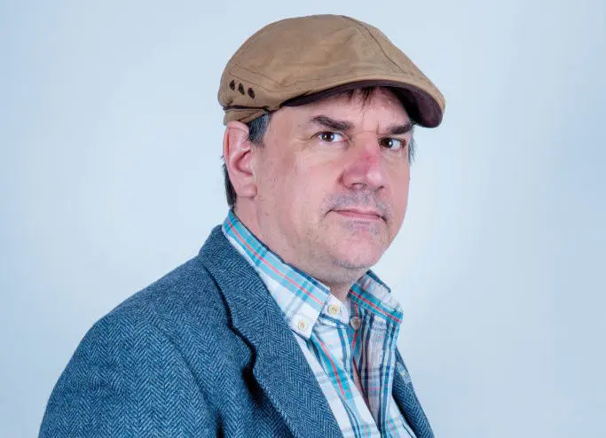 Portrait of game designer Ron Gilbert wearing a brown flat cap and a grey tweed blazer