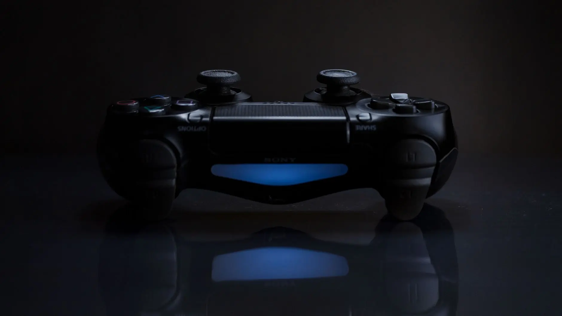 A black PlayStation DualShock 4 controller with a glowing blue light bar on a reflective surface
