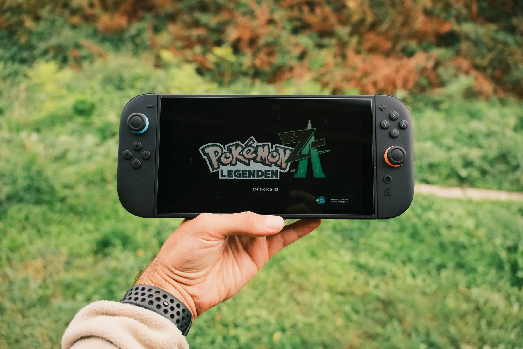 A hand holding a Nintendo Switch console displaying the title screen of Pokémon Legends: Arceus