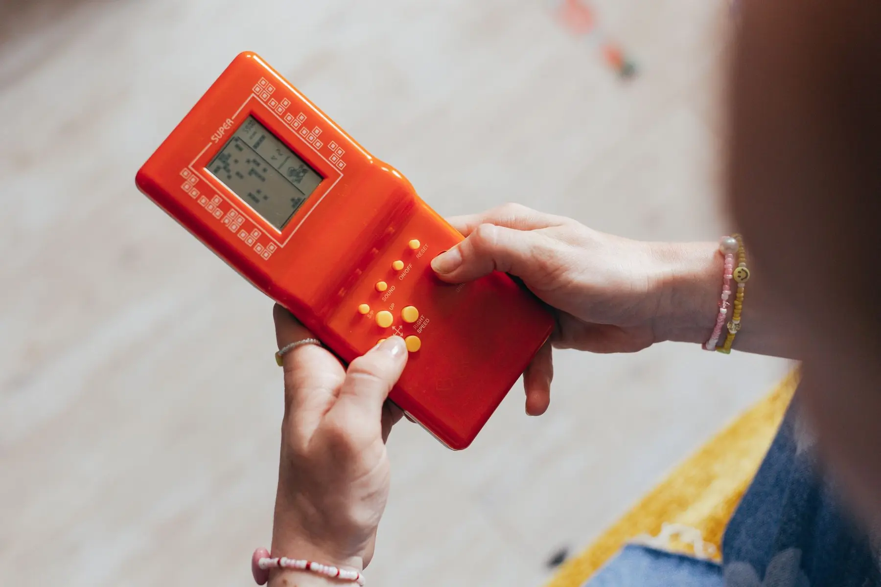 Hands holding a red handheld electronic game device with a small LCD screen showing a puzzle game